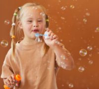 Waist up portrait of cute girl with down syndrome blowing bubbles while posing against brown background in studio, copy space